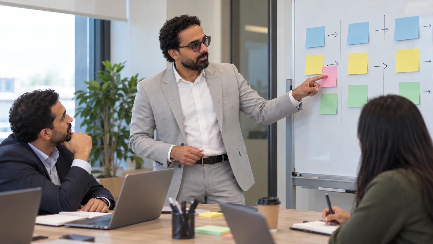 أشرف إبراهيم يشير إلى أعمدة من الملاحظات الملوّنة على السبورة، بينما يستمع زميل إلى المكتب وآخر يدوّن ملاحظات